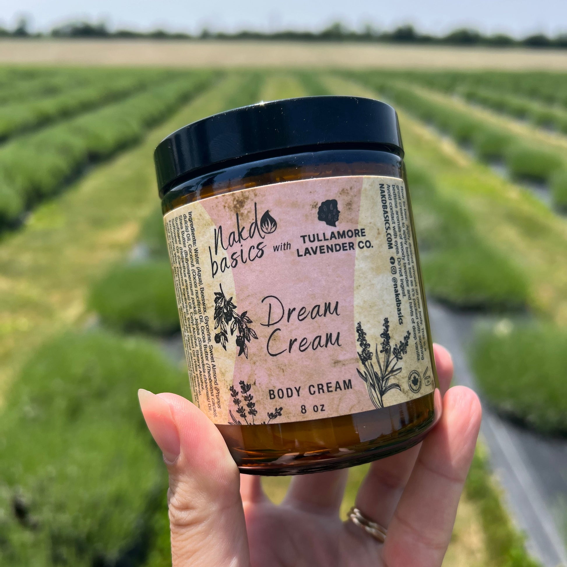 White woman's hand holds a jar with pink label that reads "Nakd Basics and Tullamore Lavender Co. Dream Cream" - green rows of plants in background.