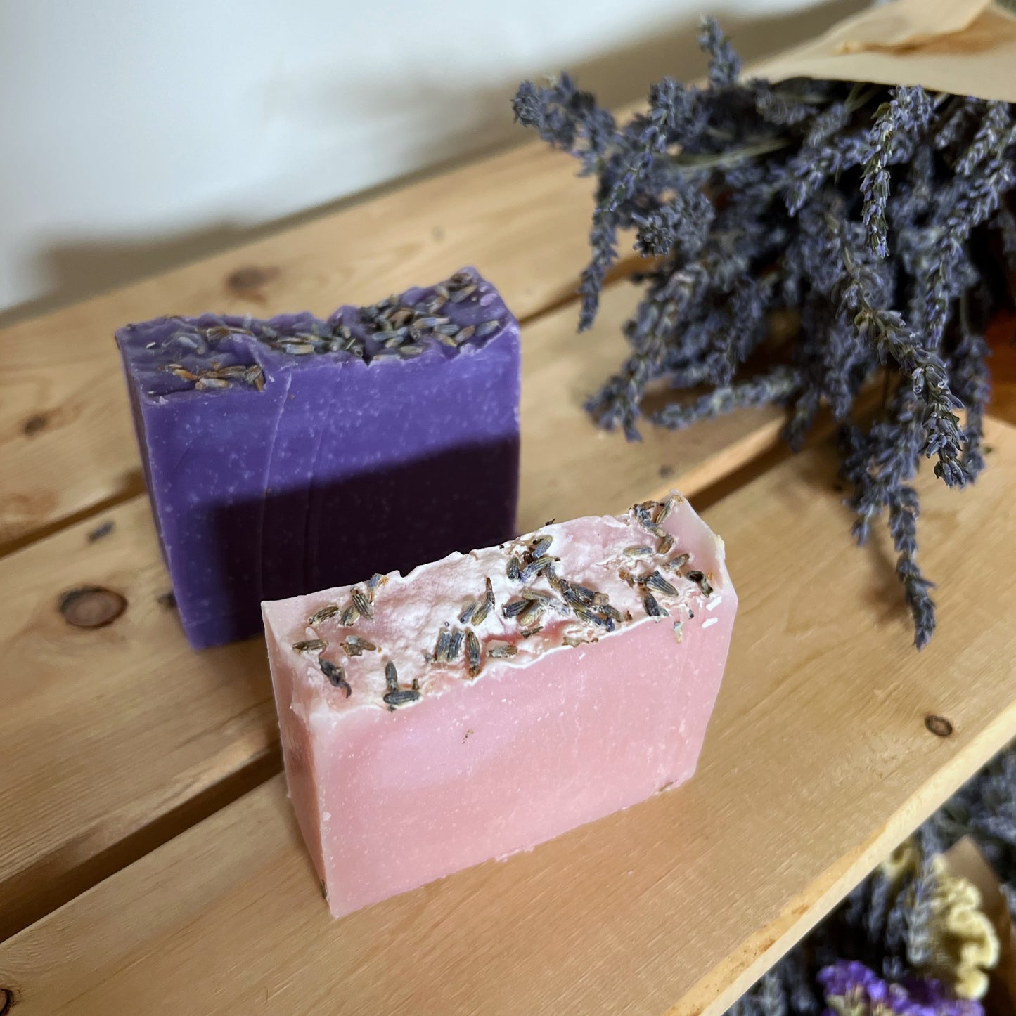 A pink bar of soap and purple bar of soap with lavender buds ontop. Lavender bouquet in background.