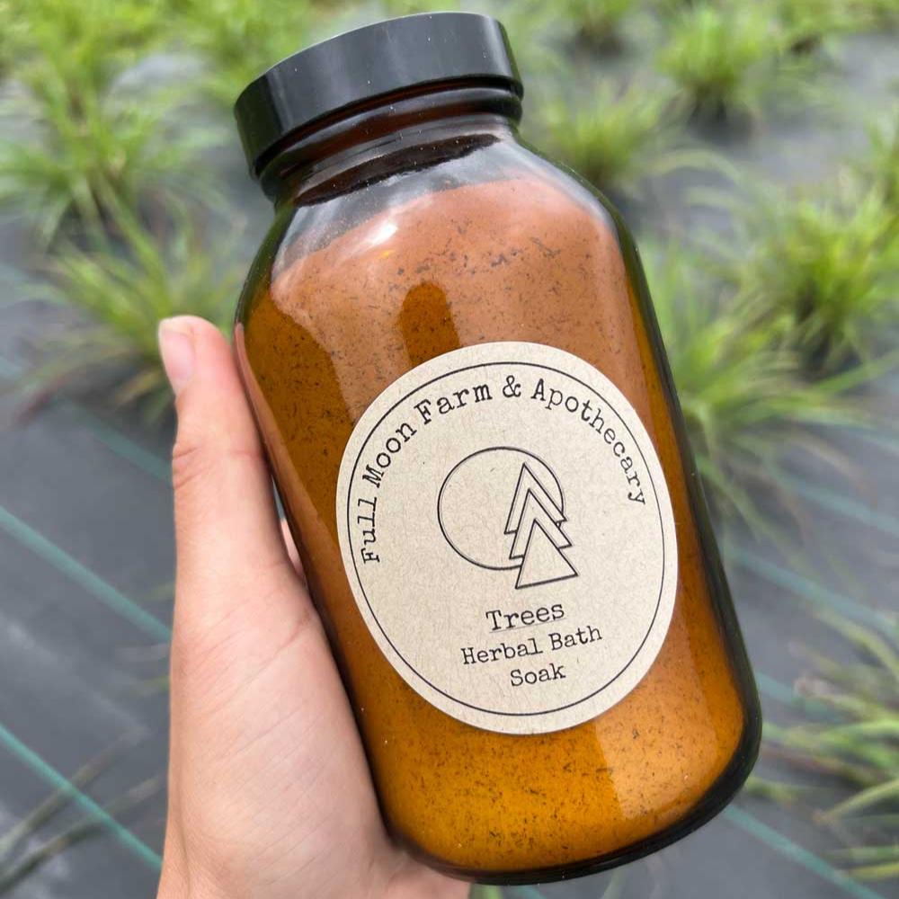 White hand holds amber jar with "Full Moon Apothecary Trees Herbal Bath Soak" label in front of green grasses growing through black weed barrier