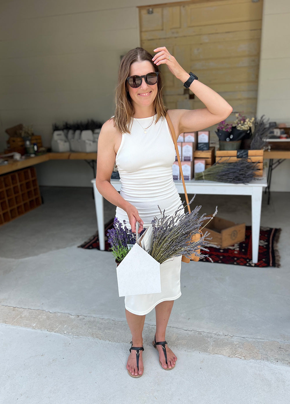 Woman in white dress and sunglasses smiles while brushing back her hair and holding a white box full of purple and green lavender.
