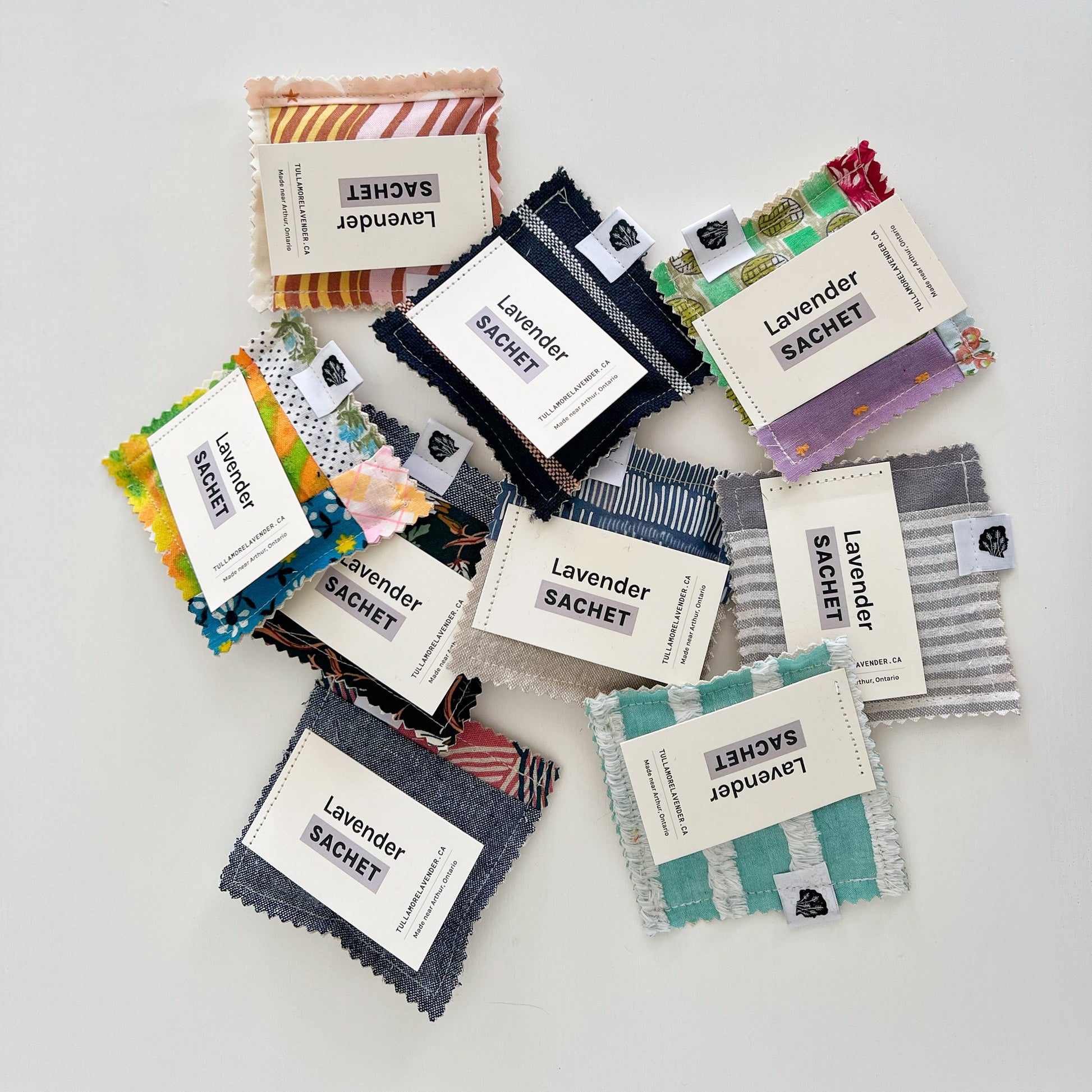 Many colourful fabric sachets lay on white background.  Tags say "Lavender Sachet"