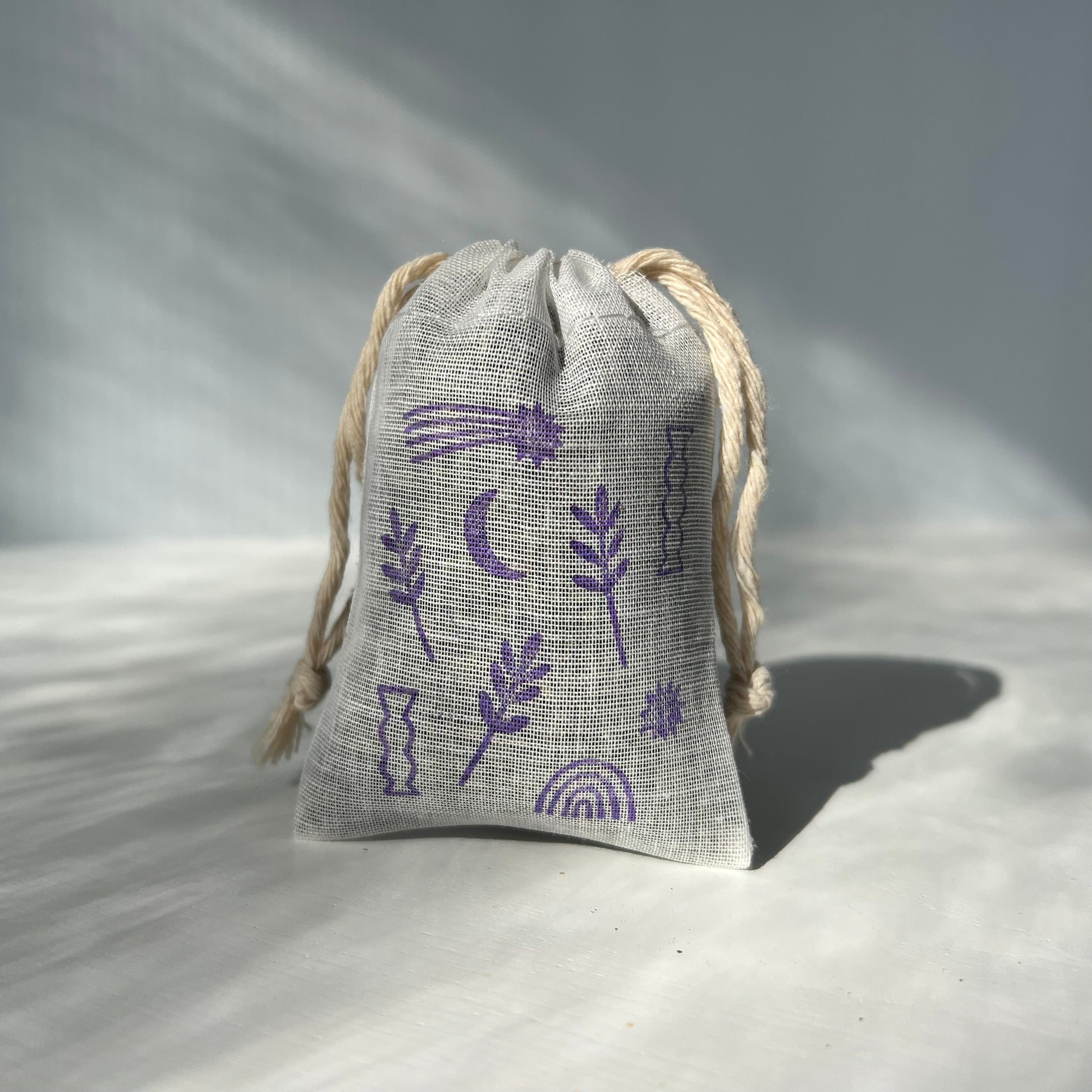 Cotton sachet with two strings hanging from the top and a purple stamped imprint of flowers, starts and rainbows.