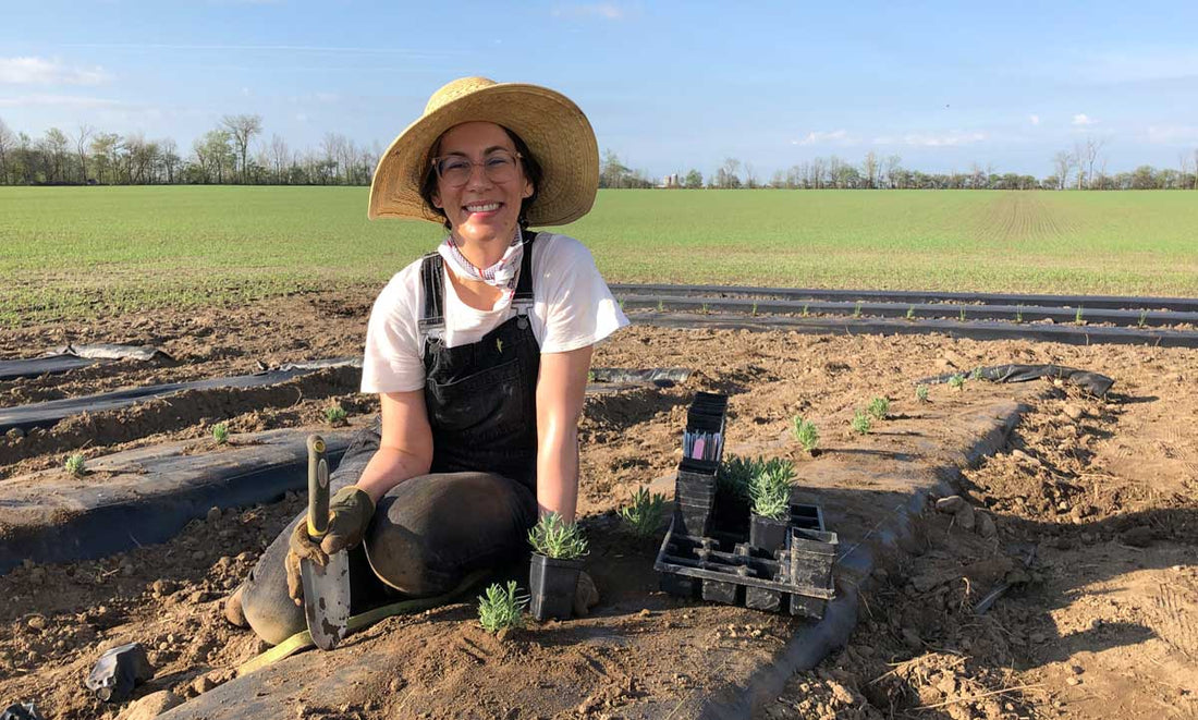 Stephanie in sun hat and overalls smile with trowel in here hand surrounded by soil and lavender plants just planted in soil