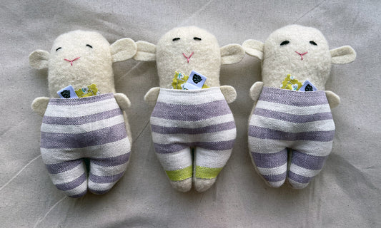 Three wool lamb stuffies lay side by side with purple striped pants on and small sachets of lavender peeking out of their pants.
