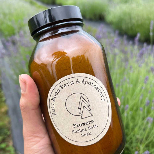 White hand holds amber jar with "Full Moon Farm & Apothecary Flowers" label in front of green and purple lavender plants