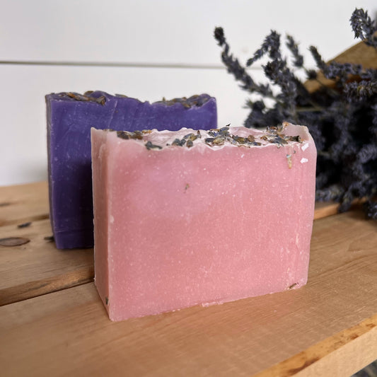 A pink bar of soap and purple bar of soap with lavender buds ontop. Lavender bouquet in background.