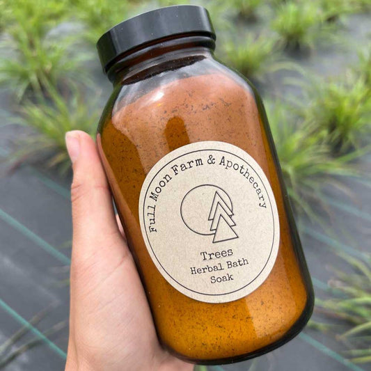 White hand holds amber jar with "Full Moon Apothecary Trees Herbal Bath Soak" label in front of green grasses growing through black weed barrier