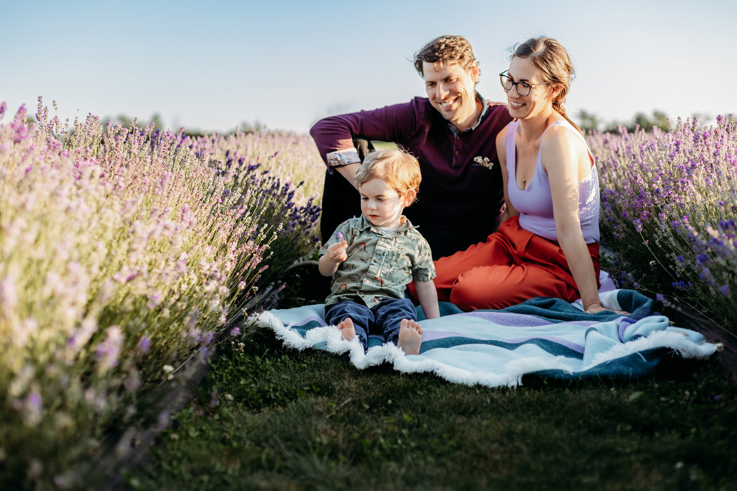 Steve, Stephanie and Felix (child) sit on purple and green blanket between two blooming rows of purple lavender.