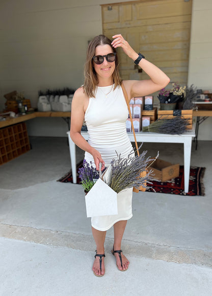 Woman in white dress and sunglasses smiles while brushing back her hair and holding a white box full of purple and green lavender.