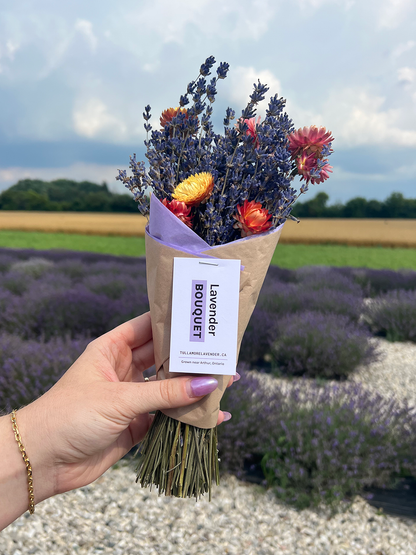 A hand holding a small dried flower bouquet with lavender, orange flowers, and a paper label reading 'Lavender Bouquet' against a blurred lavender field background.