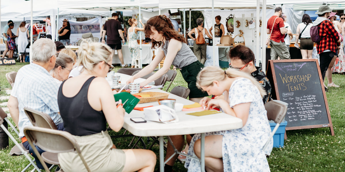 People sitting at a table with art supplies in an outdoor setting, likely at a craft fair or workshop.