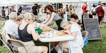 People sitting at a table with art supplies in an outdoor setting, likely at a craft fair or workshop.